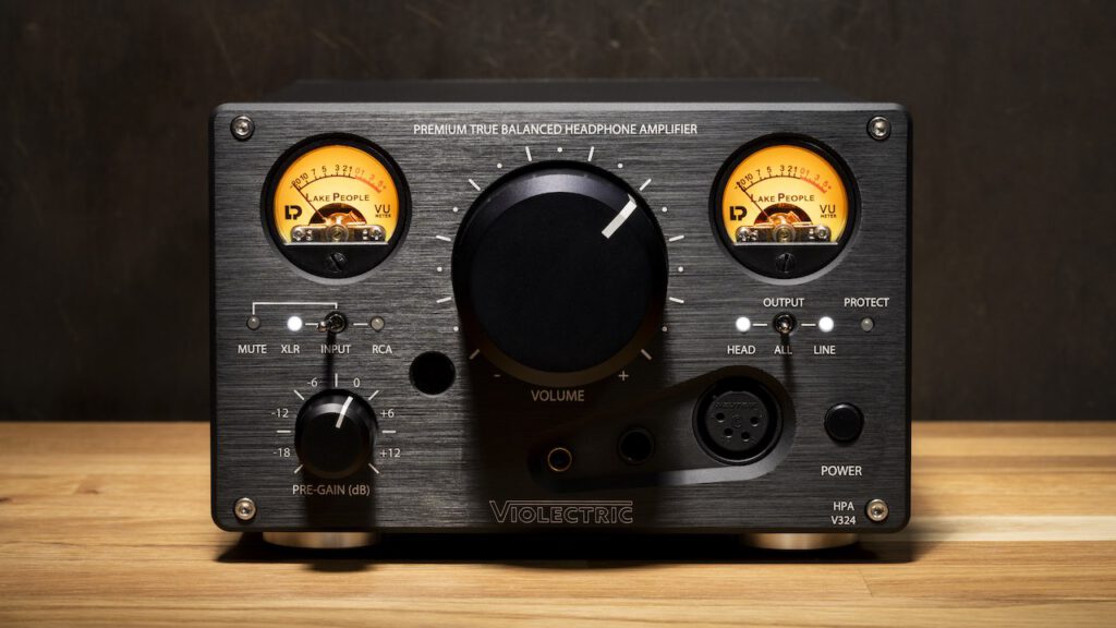 Front view of the Violectric HPA V324 headphone amplifier, featuring dual illuminated VU meters, a large central volume knob, various input/output controls, and headphone jacks, displayed on a wooden surface against a dark background.