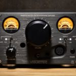 Front view of the Violectric HPA V324 headphone amplifier, featuring dual illuminated VU meters, a large central volume knob, various input/output controls, and headphone jacks, displayed on a wooden surface against a dark background.