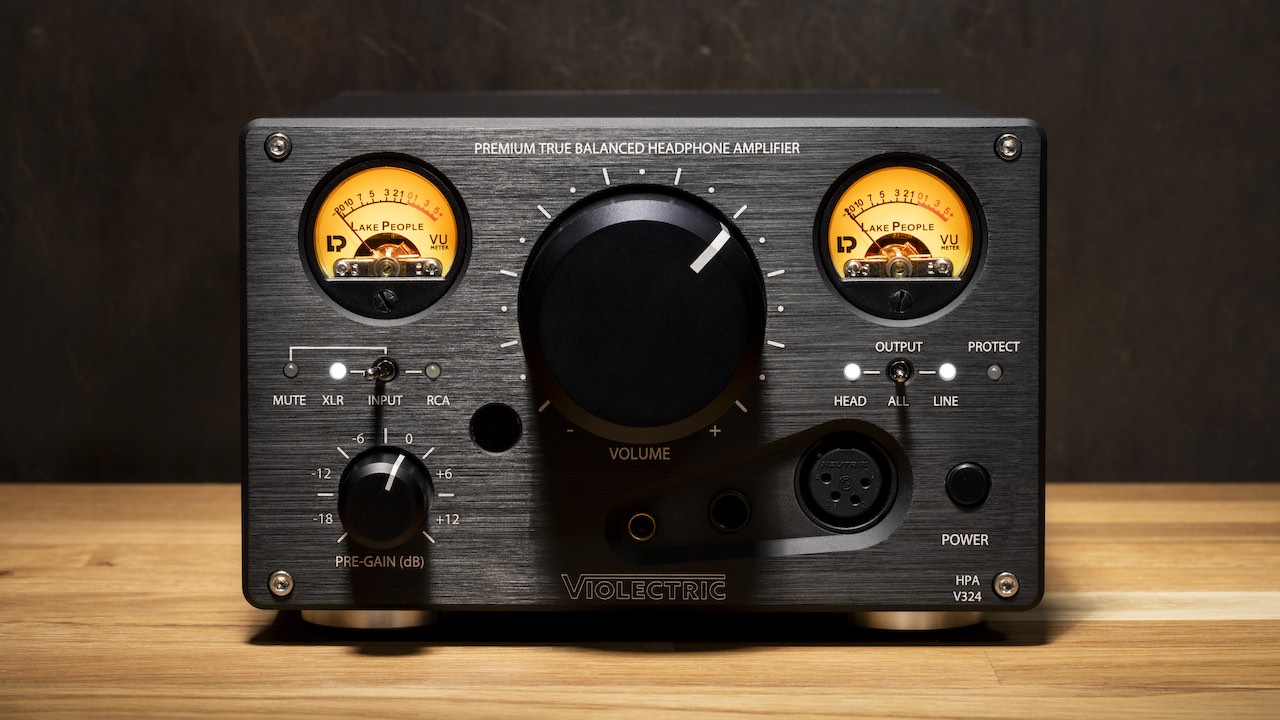 Front view of the Violectric HPA V324 headphone amplifier, featuring dual illuminated VU meters, a large central volume knob, various input/output controls, and headphone jacks, displayed on a wooden surface against a dark background.