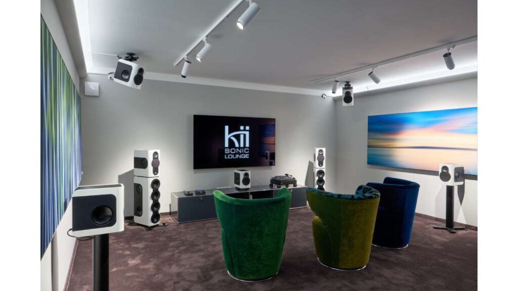 Kii Sonic Lounge demo room with a large screen and modern Dolby Atmos speaker system. Multiple speakers are placed around the room and mounted on the ceiling, with colorful armchairs in front for visitors.