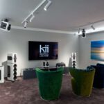 Kii Sonic Lounge demo room with a large screen and modern Dolby Atmos speaker system. Multiple speakers are placed around the room and mounted on the ceiling, with colorful armchairs in front for visitors.