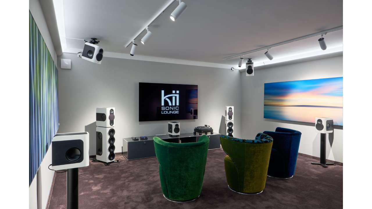 Kii Sonic Lounge demo room with a large screen and modern Dolby Atmos speaker system. Multiple speakers are placed around the room and mounted on the ceiling, with colorful armchairs in front for visitors.