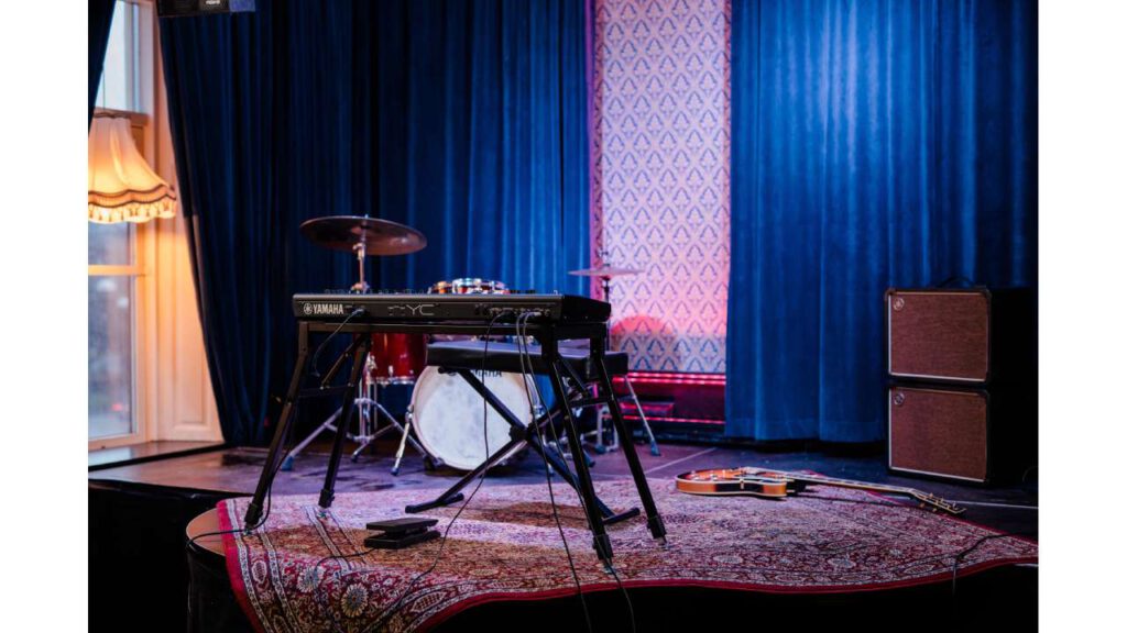 Stage with a Yamaha YC stage keyboard on a stand, a drum set in the background, an amplifier on the right side, and a guitar lying on the carpet in front of the keyboard.