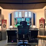 Professional recording studio with a large mixing console, central display and ergonomic studio chair, surrounded by multiple large PSI Audio floor-standing speakers. Acoustic panels on the ceiling and walls optimize the room’s sound treatment.