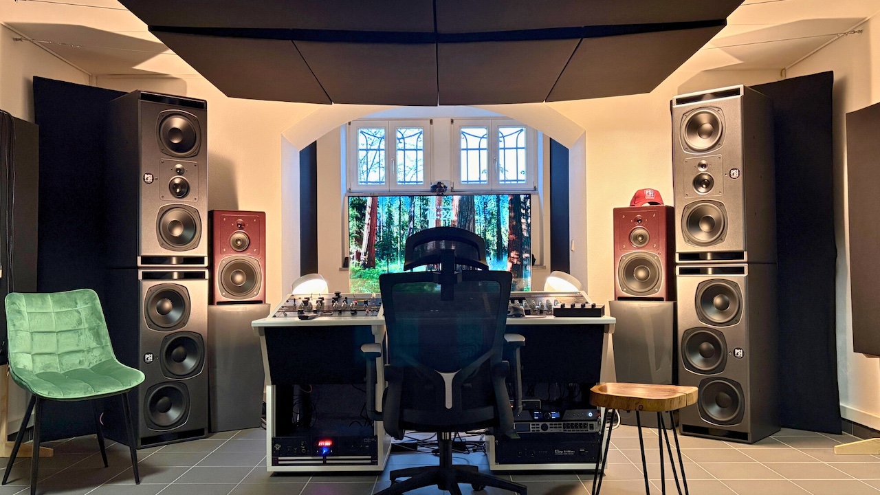 Professional recording studio with a large mixing console, central display and ergonomic studio chair, surrounded by multiple large PSI Audio floor-standing speakers. Acoustic panels on the ceiling and walls optimize the room’s sound treatment.