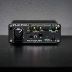 Lake People MC10 monitor controller on a studio desk, featuring a volume knob, headphone jack, and input/output status LEDs.
