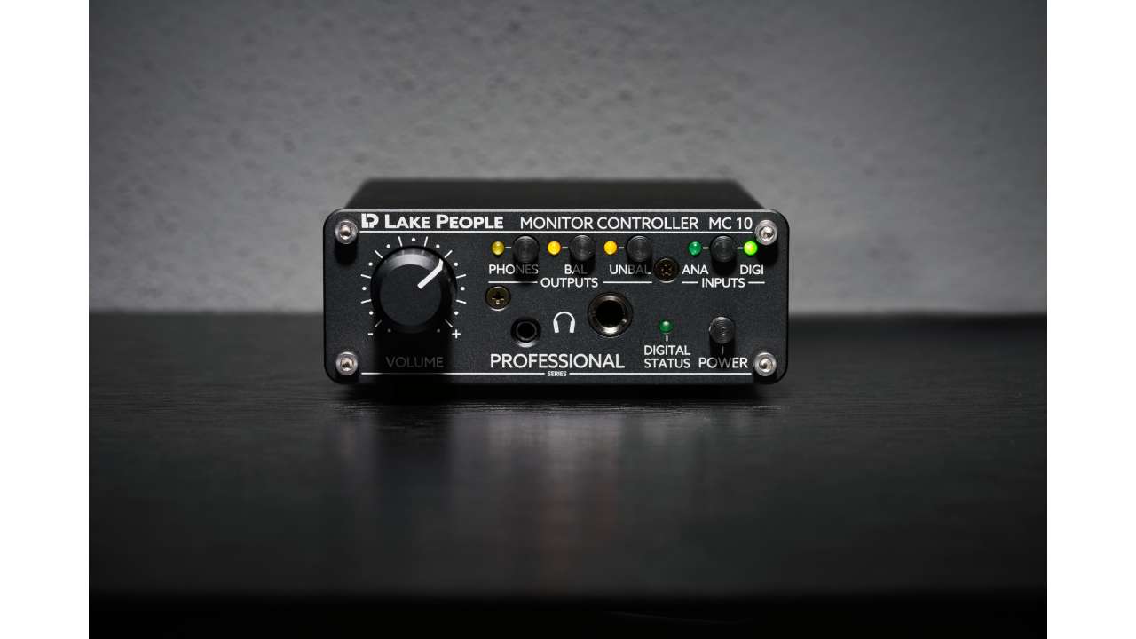 Lake People MC10 monitor controller on a studio desk, featuring a volume knob, headphone jack, and input/output status LEDs.