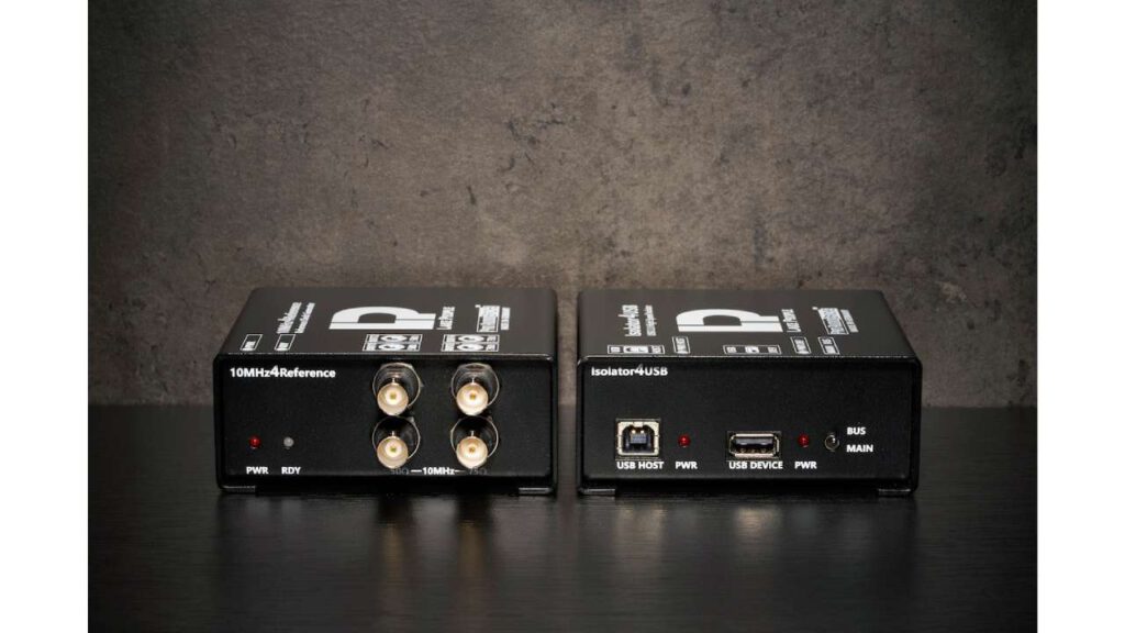 Two compact Lake People pro audio devices on a table: a 10MHz reference clock generator and a USB isolator with ports and status LEDs in a studio setting.